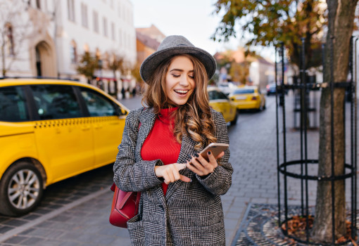Good-looking business woman texting message while walking on the street. Refined lady in elegant coat looking at phone screen, standing on avenue with taxi on background.. Good-looking business woman texting message while walking on the street. Refined lady in elegant coat looking at phone screen, standing on avenue with taxi on background.